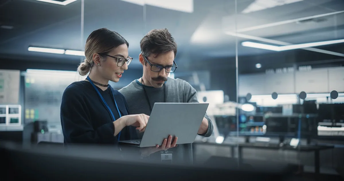 Two IT professionals in a high-tech control room discuss data on a laptop, representing collaboration and decision-making in technology.